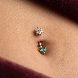 May include: A belly button piercing featuring a gold-coloured bar with two green, teardrop-shaped gems. Above the piercing is a small, flower-shaped stud with iridescent stones. The jewellery is set against skin.