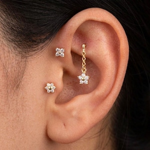 May include: Close-up of an ear adorned with three pieces of jewelry. A small, gold flower stud is visible, along with a square stud with four clear stones. A gold chain suspends a flower-shaped earring with clear stones.