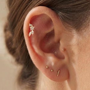 May include: A gold ear cartilage stud featuring a flower design with clear stones, accompanied by two small gold ball studs and a thin gold bar stud in the earlobe. The jewellery pieces are metallic gold with clear, faceted stones set in the flower.