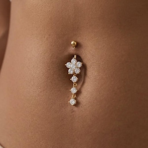 Könnte beinhalten: Goldfarbener Bauchnabelpiercing mit einem Blumendesign und hängenden, klaren Edelsteinen. Der obere Ball und die Blume sind fixiert, während die Edelsteine frei hängen. Der Schmuck ist für ein Bauchnabelpiercing konzipiert.