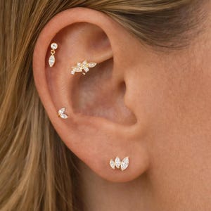 Puede incluir: Primer plano de una oreja adornada con múltiples pendientes dorados. Los pendientes presentan gemas claras, de talla marquesa y redonda. Un pendiente tiene forma de lágrima colgante, mientras que otros son tachuelas y una barra curva.