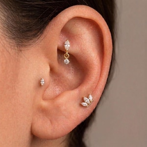 Puede incluir: Tres pendientes dorados con piedras transparentes de talla marquesa. Un pendiente es un dije colgante en el rook, otro es un pequeño perno en el trago y otro es un perno de racimo con varias piedras en el lóbulo, todos con monturas doradas.
