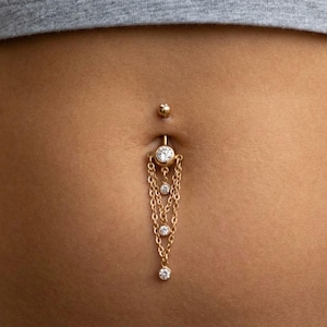 May include: Gold belly button ring with a dangling chain design. The ring features a small ball on top, a large clear gem at the centre, and a series of chains with smaller gems. The jewellery is designed to be worn on the navel.