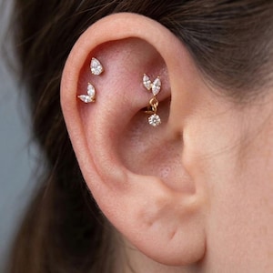 Può includere: Un set di piercing per orecchie in oro composto da tre pezzi distinti. Due borchie hanno la forma di gocce e foglie marquise, mentre il terzo pezzo è un cerchio d'oro con una foglia marquise pendente e una piccola pietra rotonda trasparente.