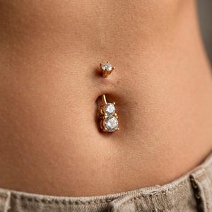 Pode incluir: Close-up de um piercing no umbigo com duas pedras preciosas redondas e brilhantes cravejadas em ouro. Um pequeno pino de ouro correspondente está acima do umbigo. A pele tem um tom quente e neutro.