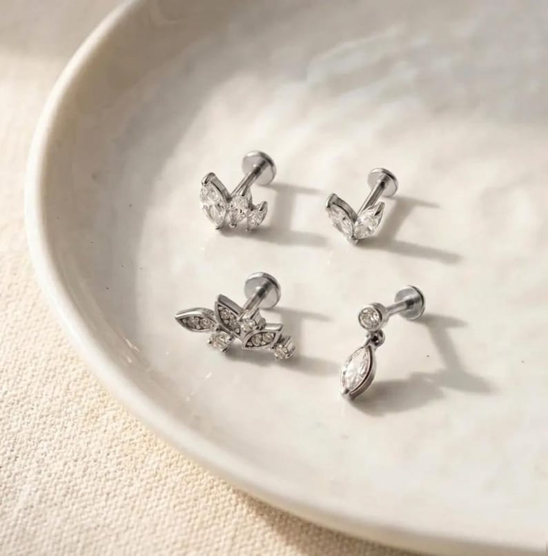 Puede incluir: Cuatro piezas de joyer&iacute;a plateada con piedras preciosas claras y brillantes. Dos piezas presentan un dise&ntilde;o en forma de hoja, una con forma de l&aacute;grima colgante, y la &uacute;ltima tiene un grupo de piedras. La joyer&iacute;a se exhibe en un plato blanco poco profundo.