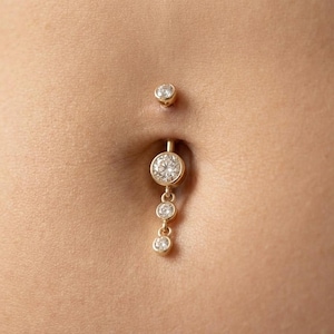 May include: Gold-coloured belly button piercing with three dangling clear gemstones. The top stud is a small, single gemstone. The main charm features a larger, round gemstone with two smaller gems below.