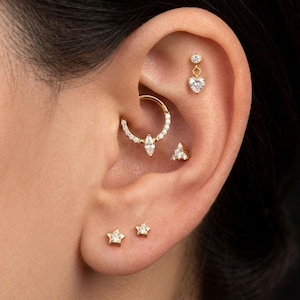 Pode incluir: Close-up de uma orelha adornada com vários brincos dourados. Um brinco pendurado em forma de coração, um brinco de triângulo e dois brincos de estrela são visíveis. Um brinco de argola dourado com uma pedra central em forma de marquesa e gemas circundantes também está presente.