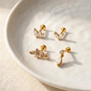 Puede incluir: Cuatro pendientes dorados con gemas claras y brillantes dispuestas en varios dise&ntilde;os florales y de hojas. Los pendientes se exhiben en un plato de cer&aacute;mica blanca, mostrando sus intrincados detalles y su estilo elegante.