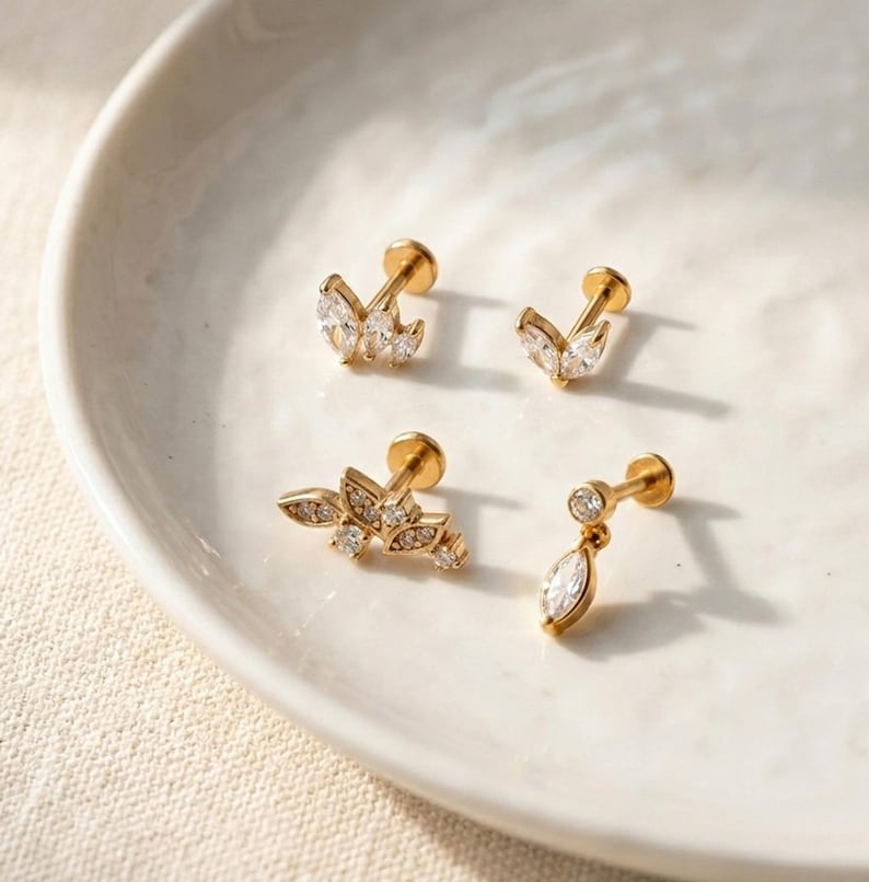 Puede incluir: Cuatro pendientes dorados con gemas claras y brillantes dispuestas en varios dise&ntilde;os florales y de hojas. Los pendientes se exhiben en un plato de cer&aacute;mica blanca, mostrando sus intrincados detalles y su estilo elegante.