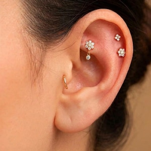 May include: A gold hoop earring in the tragus and three gold stud earrings in the ear cartilage. The cartilage studs feature clear, sparkling stones arranged in floral shapes, with one stud dangling a small, round, clear stone from its center.