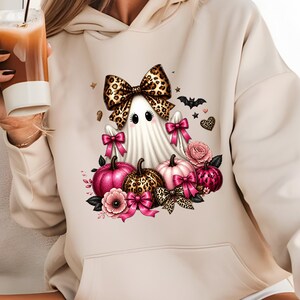 Könnte beinhalten: Ein beigefarbener Kapuzenpullover mit einem Grafikdruck eines weißen Geistes, der eine große Schleife mit Leopardenmuster trägt. Das Design umfasst rosa Kürbisse, Kürbisse mit Leopardenmuster, rosa Blumen, leuchtend rosa Schleifen, eine kleine schwarze Fledermaus und Sterne.