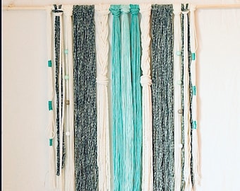 May include: A wall hanging made of white, gray, and teal yarn with wooden dowel at the top. The yarn is tied in knots and hangs in strands of different lengths.