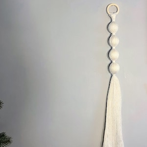 May include: White macrame wall hanging with a wooden ring at the top and four white yarn balls hanging from it.