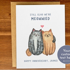 May include: A white card with a watercolour illustration of two cats, one grey and one orange, with a heart above them. The text reads "Still glad we're Meowwied" and "Happy Anniversary, Jaimie!"