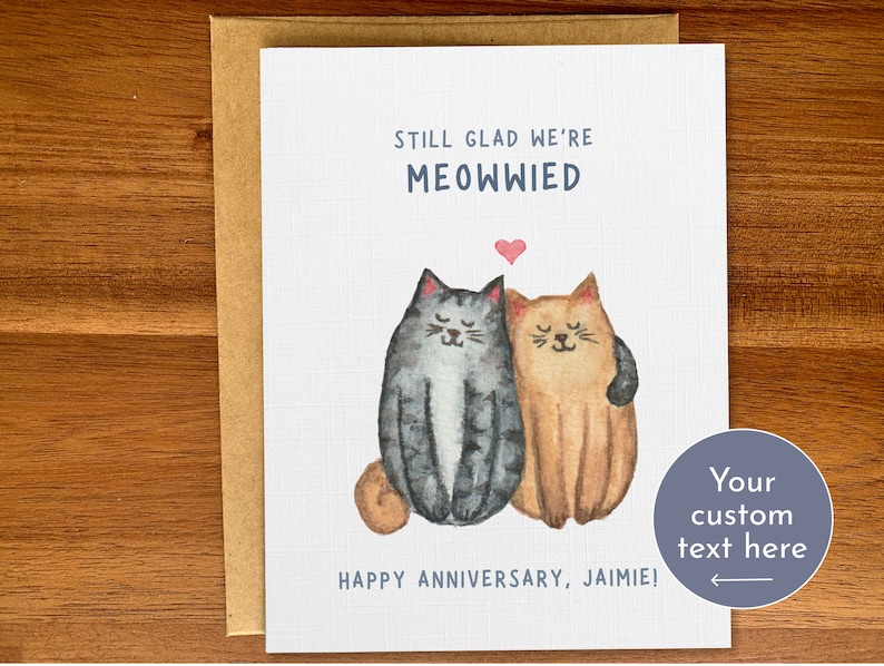 May include: A white card with a watercolour illustration of two cats, one grey and one orange, with a heart above them. The text reads "Still glad we're Meowwied" and "Happy Anniversary, Jaimie!"