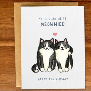 May include: A greeting card with a black and white illustration of two cats with a pink heart above them. The text reads "Still glad we're meowwied. Happy Anniversary."