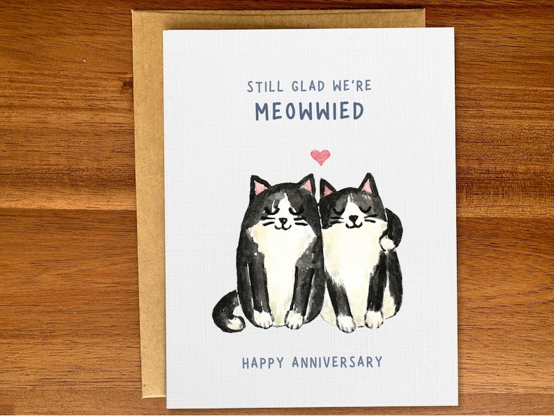 May include: A greeting card with a black and white illustration of two cats with a pink heart above them. The text reads "Still glad we're meowwied. Happy Anniversary."