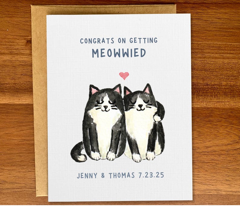 May include: A white card with a black and white illustration of two cats with a pink heart above them. The text on the card reads "Congrats on getting Meowwied" and "Jenny & Thomas 7.23.25".