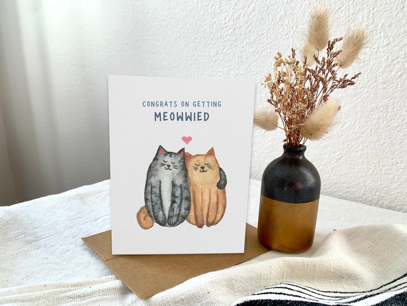 May include: A white card with the text "Congrats on getting Meowwied" and a watercolor illustration of two cats hugging. The cat on the left is gray and white, and the cat on the right is orange. A small pink heart is above the cats.