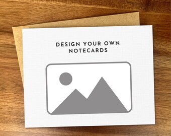 May include: A white notecard with the text "DESIGN YOUR OWN NOTECARDS" above a gray graphic of a mountain range and a circle. The notecard is on top of a brown envelope, all on a wooden surface.