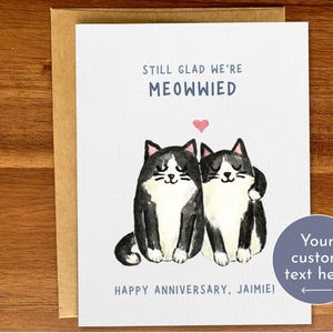 May include: A greeting card featuring a black and white illustration of two cats with a heart above them. The text on the card reads "Still glad we're Meowwied" and "Happy Anniversary, Jamie!" with a space for custom text.