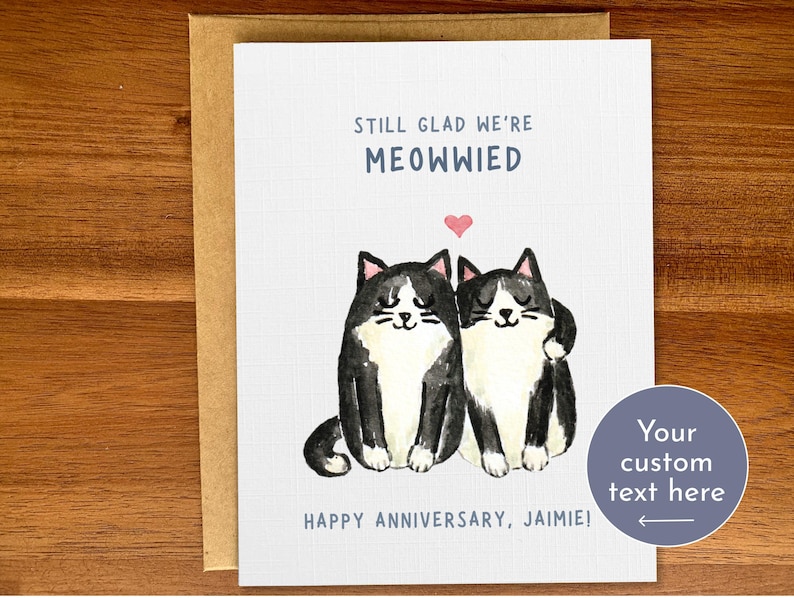 May include: A greeting card featuring a black and white illustration of two cats with a heart above them. The text on the card reads "Still glad we're Meowwied" and "Happy Anniversary, Jamie!" with a space for custom text.