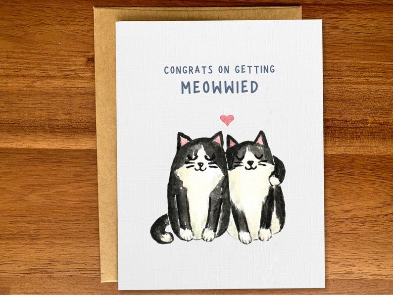 May include: A greeting card with a black and white illustration of two cats with a heart above them. The text on the card reads "Congrats on getting Meowwied".