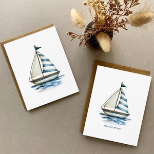 May include: Two watercolour greeting cards with brown borders. Each card features a sailboat with blue and white striped sails on blue water. One card has the text "YOU FLOAT MY BOAT".