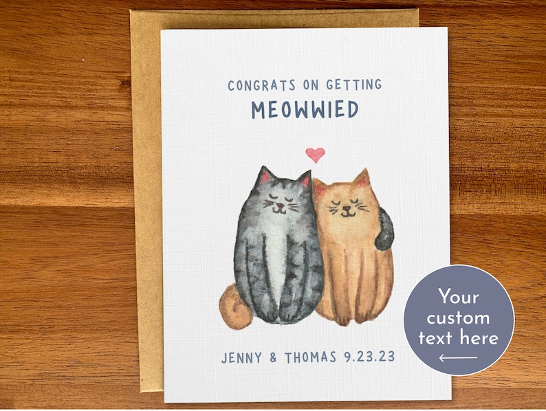 May include: A white card with a watercolor illustration of two cats, one gray and one orange, with a heart above them. The text on the card reads "Congrats on getting Meowwied" and "Jenny & Thomas 9.23.23".