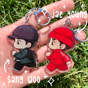 Peut inclure: Deux porte-clés de personnages de dessins animés, l'un en rouge et l'autre en vert, avec les noms "Jae Young" et "Sang Woo" écrits sur l'image.