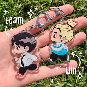 May include: Two cartoon character keychains with a silver keychain attached. The keychain on the left is a black-haired character wearing a white shirt and black pants. The keychain on the right is a blonde character wearing a blue jacket and black pants. The text "team" is written on the left side of the image and the text "win" is written on the right side of the image.