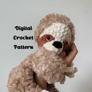 May include: A crocheted sloth amigurumi toy with brown and white fur. The sloth has a brown nose and eyes, and a pink mouth.
