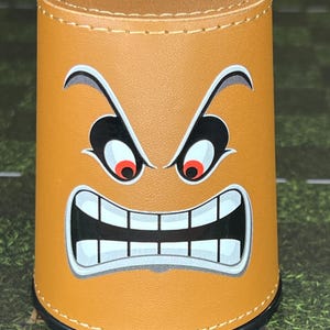 May include: A cylindrical brown dice cup with a cartoon angry face design. The face features white teeth, red eyes, and black eyebrows. The cup has a black base and a stitched top edge.