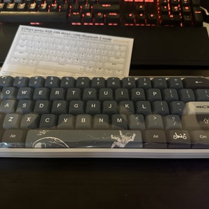 May include: A compact, grey and blue mechanical keyboard with white lettering, packaged in plastic. The keyboard has a space-themed design on the space bar and other keys. A paper with keyboard layout is behind it.