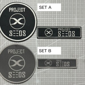 Op de afbeelding: Twee sets geborduurde patches met het Project Seeds logo. Set A bevat een ronde patch met een zwarte achtergrond en witte tekst en logo, en een rechthoekige patch met een zwarte achtergrond en witte tekst en logo. Set B bevat een ronde patch met een grijze achtergrond en witte tekst en logo, en een rechthoekige patch met een grijze achtergrond en witte tekst en logo.