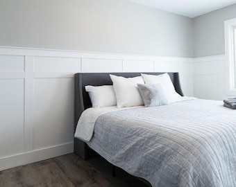May include: A gray upholstered bed frame with a white headboard and white bedding. The bed is in a bedroom with white wainscoting on the walls.