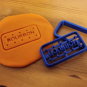 May include: Blue plastic cookie cutter with the word "BOURBON" embossed on it. The cutter is on top of orange modelling clay with the word "BOURBON" imprinted in it.