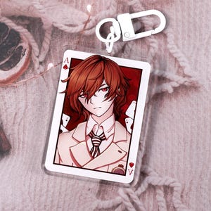 Peut inclure: Porte-clés en acrylique transparent en forme de carte à jouer, orné d'un personnage de style anime. Le personnage a les cheveux bruns et porte un costume. Le porte-clés est fixé à un fermoir argenté. Fond texturé rose clair.