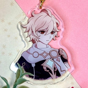 Puede incluir: Un llavero de acrílico transparente con un personaje de anime de cabello rubio, que lleva un atuendo blanco y negro con detalles dorados. El personaje tiene ojos rojos y una expresión seria.