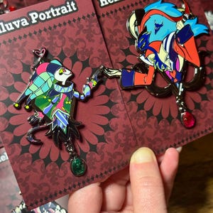 Peut inclure: Deux &eacute;pingles &eacute;maill&eacute;es repr&eacute;sentant des personnages de "Helluva Boss". L'une repr&eacute;sente un personnage de type bouffon aux couleurs vives, l'autre un personnage aux cheveux bleus et aux accents rouges. Les deux &eacute;pingles ont des breloques en forme de pierres pr&eacute;cieuses et sont sur des cartes marron.