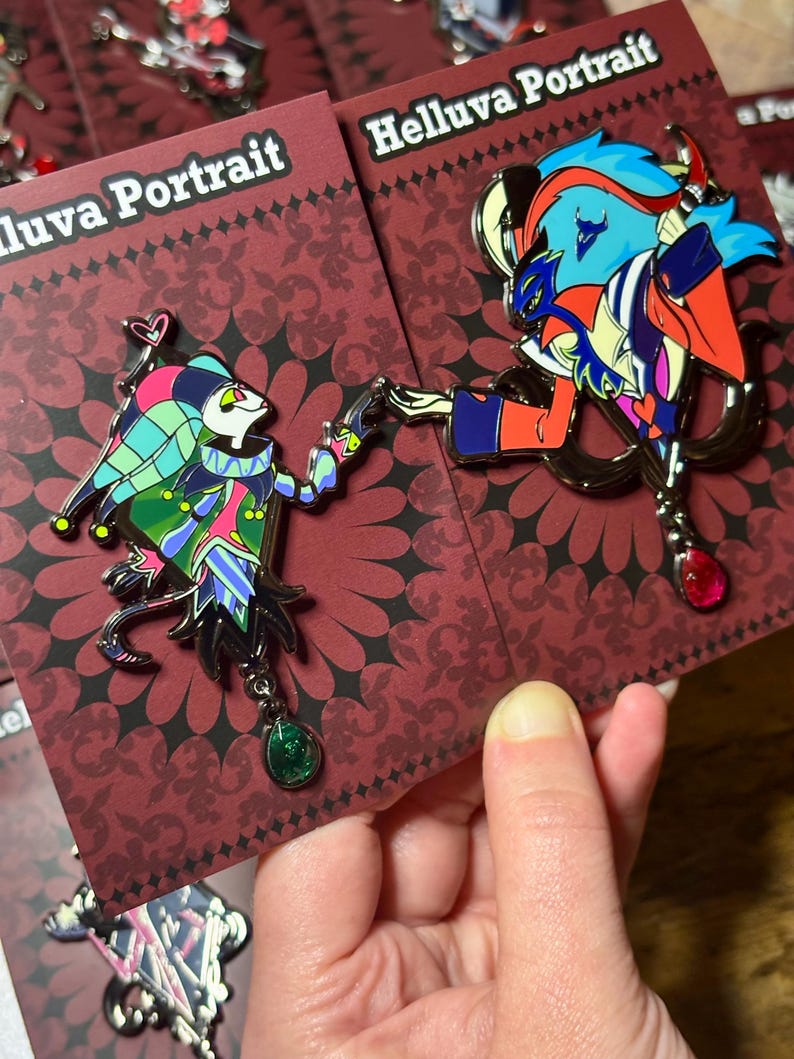 Peut inclure: Deux &eacute;pingles &eacute;maill&eacute;es repr&eacute;sentant des personnages de "Helluva Boss". L'une repr&eacute;sente un personnage de type bouffon aux couleurs vives, l'autre un personnage aux cheveux bleus et aux accents rouges. Les deux &eacute;pingles ont des breloques en forme de pierres pr&eacute;cieuses et sont sur des cartes marron.
