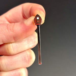 May include: A silver-toned enamel pin shaped like a stick with a red and white flame at the top.