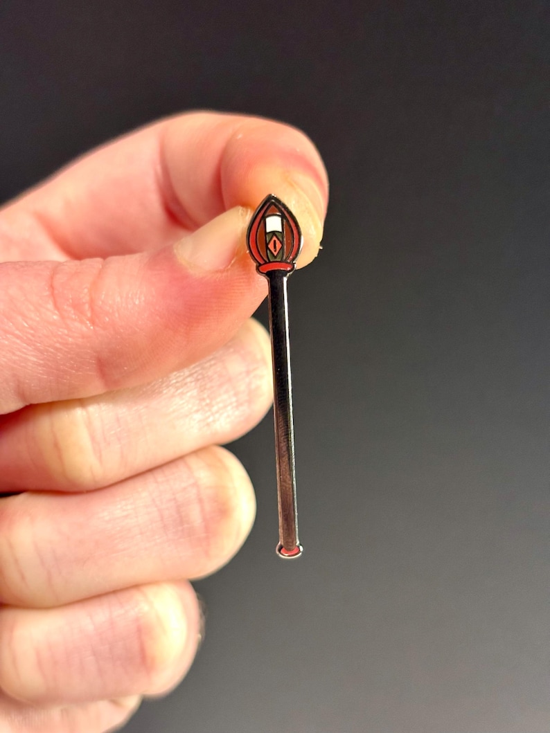 May include: A silver-toned enamel pin shaped like a stick with a red and white flame at the top.