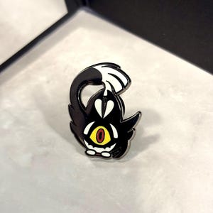 May include: Black and white enamel pin of a cartoon character with a single yellow eye and red pupils. The character has a black body and white fur on its head and tail. The character is curled up with its tail over its head.