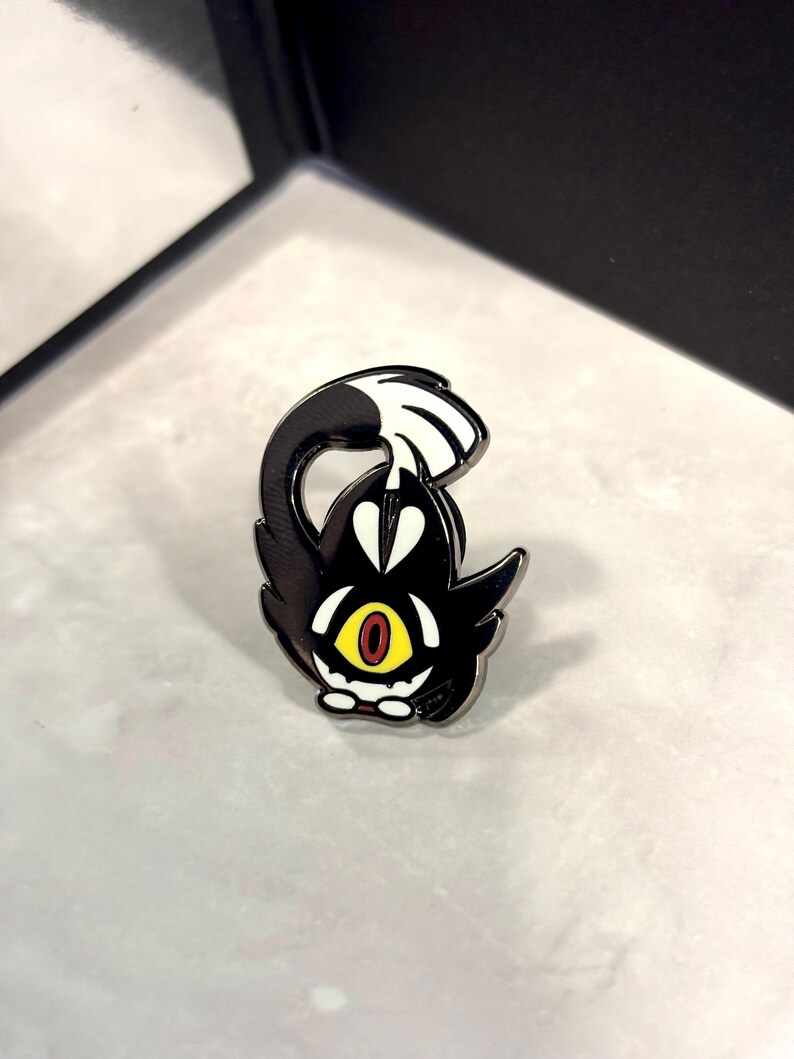 May include: Black and white enamel pin of a cartoon character with a single yellow eye and red pupils. The character has a black body and white fur on its head and tail. The character is curled up with its tail over its head.