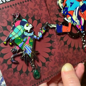 Peut inclure: Deux &eacute;pingles &eacute;maill&eacute;es repr&eacute;sentant des personnages de dessins anim&eacute;s color&eacute;s. L'une repr&eacute;sente un bouffon avec un pendentif en pierre verte, l'autre un personnage avec des accents bleus et rouges. Les &eacute;pingles sont sur un fond rouge &agrave; motifs avec le texte "Portrait" et "Helluva".