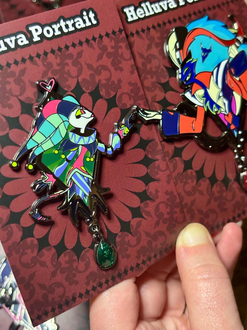 Peut inclure: Deux &eacute;pingles &eacute;maill&eacute;es repr&eacute;sentant des personnages de dessins anim&eacute;s color&eacute;s. L'une repr&eacute;sente un bouffon avec un pendentif en pierre verte, l'autre un personnage avec des accents bleus et rouges. Les &eacute;pingles sont sur un fond rouge &agrave; motifs avec le texte "Portrait" et "Helluva".