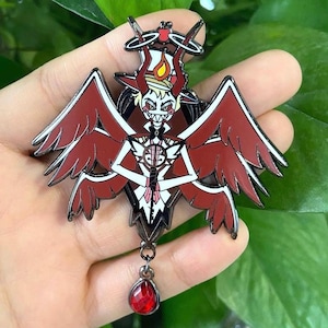 Puede incluir: Un pin de esmalte detallado que presenta un personaje de demonio estilizado con alas rojas, cuernos y un colgante de gema roja. El pin tiene un contorno negro y detalles en blanco.