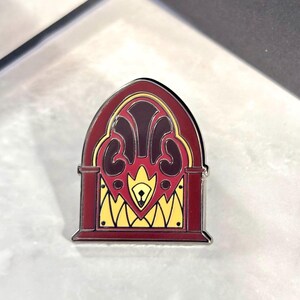May include: A red, yellow, and black enamel pin shaped like a vintage radio. The radio has a decorative design on the front with a small, black knob.